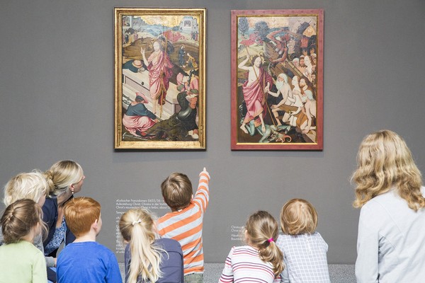 Rückansicht von einigen Kindern und zwei erwachsenen Personen, die sich zwei Gemälde angucken. Ein Kind zeigt mit dem Finger auf eines der Bilder.